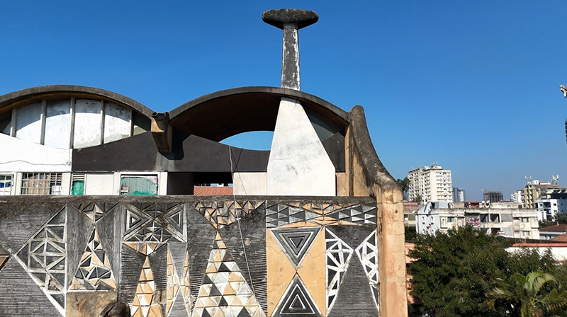  A modernist building with curved roofs, geometric facade patterns, and a distinctive chimney, set against a blue sky and an urban skyline in the background.