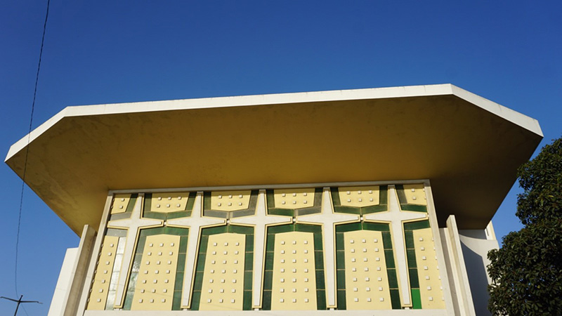  A modernist church with a large overhanging roof, geometric facade, and cross-shaped window patterns, set against a clear blue sky with trees nearby.