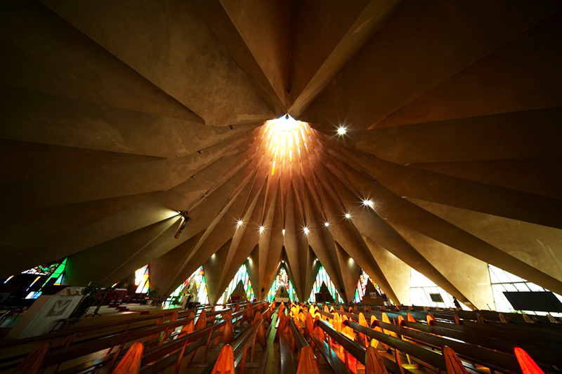 A worship space interior with a dramatic radial ceiling converging at a skylight, stained glass windows, and wooden pews illuminated by warm light.