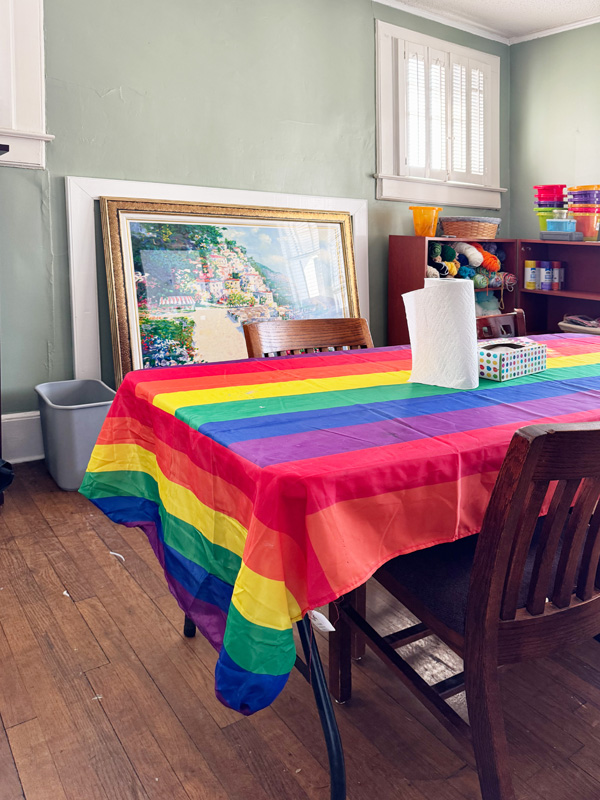 Folding table with metal legs covered by a rainbow tablecloth. A shelf in the background holds art supplies such as yarn and tulle. 