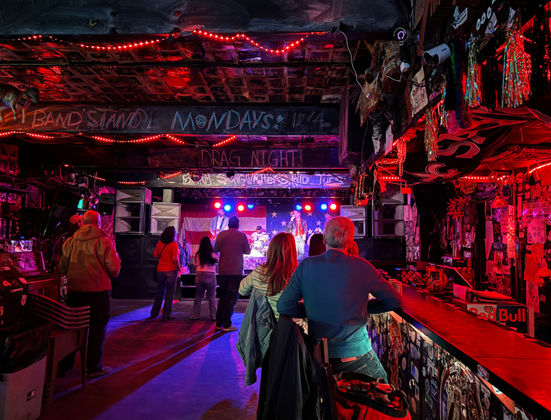 Interior of a bar with tinted red lighting coming from behind the bar.  Patrons take in a musical performance from the stage at the far end of the room.