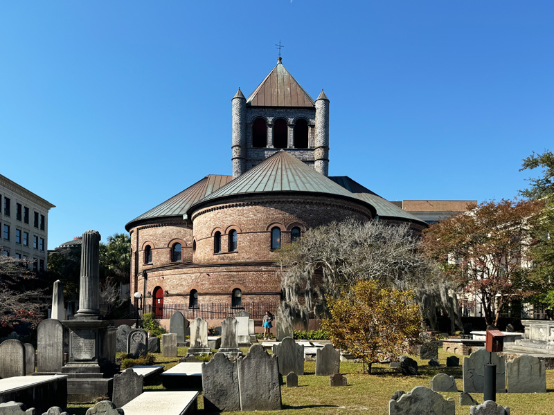  A historic round brick church with a copper roof and unique cylindrical towers, surrounded by an old graveyard with weathered tombstones. The scene is bathed in bright daylight under a clear blue sky.