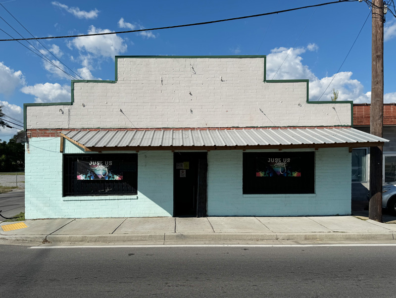 External view of cinderblock building with a glass dor flanked by large glass windows in the walls to either side. A metal corrugated roof hangs over the entrance. 