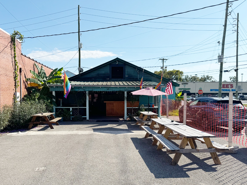 In the back patio of a restaurant seen in the background, wooden picnic benches sit atop an asphalt surface. One supports a shade umbrella. Pride flags fly from the restaurant back porch.