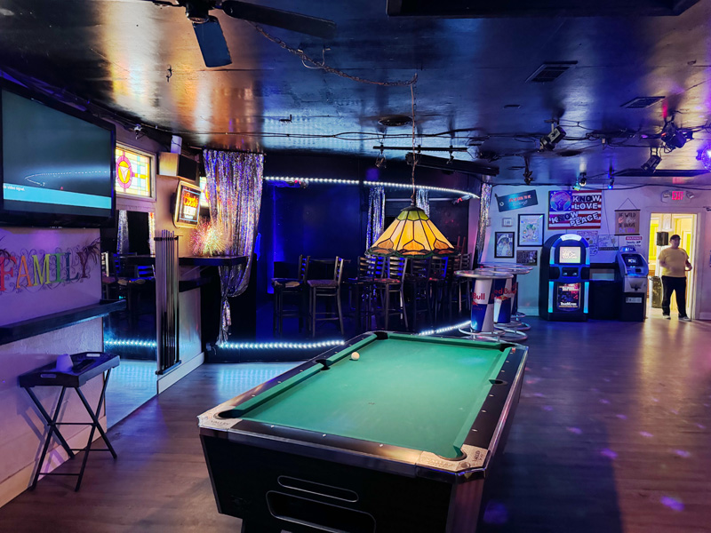 In the darkened interior of a bar, a glass pendant lamp illuminates a billiards table in the foreground. Small points of light glint on the floor behind it from a disco ball hanging somewhere out of shot.