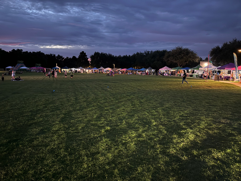 Between an open grass field and a clouded sunset sky, vendor awnings dot the horizon below a line of trees. Patrons walk between them. 