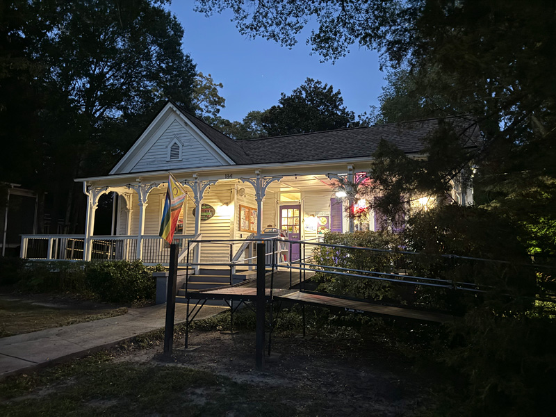 A warmly lit, historic-style bookstore with a charming wraparound porch, surrounded by trees and greenery, illuminated at dusk. A prominent Pride flag and an accessible ramp enhance its welcoming atmosphere.