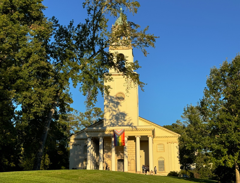 Façade of a church with a pride flag hanging between the front columns of the classical-style building. 