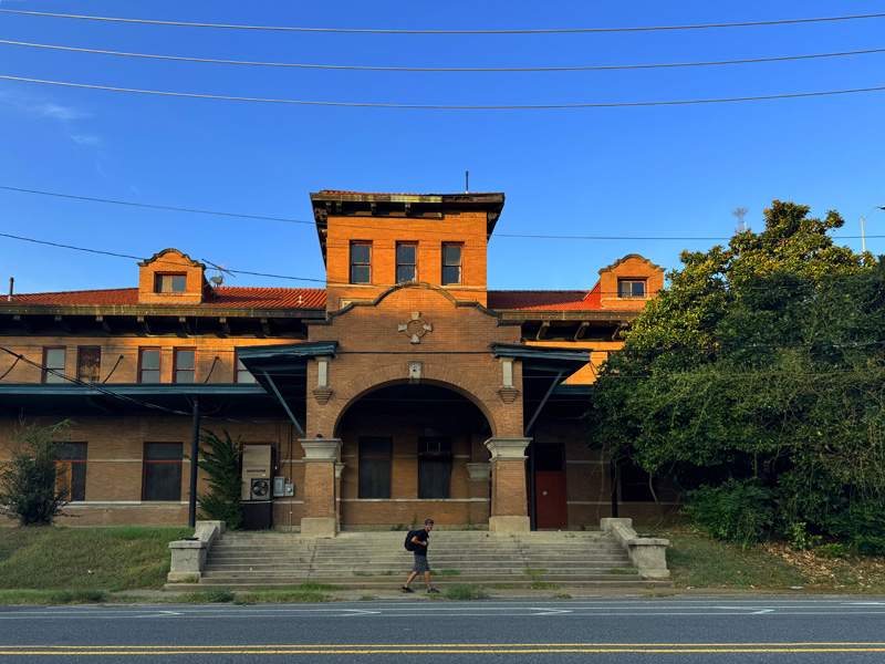 The brick building features a central arched entrance with decorative stonework and tall windows above. The roof is covered in red tiles, and smaller gable structures are visible on either side. 