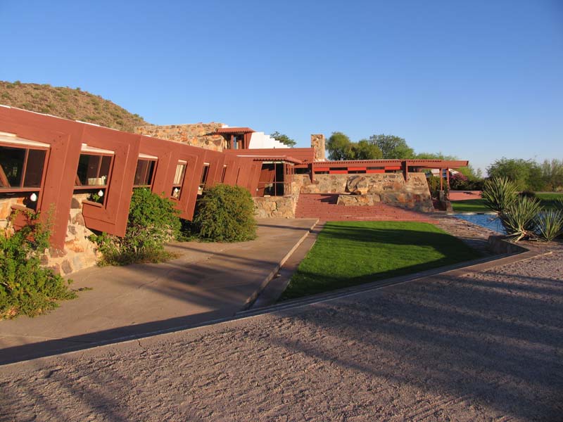 low stone and wood house with slightly canted walls and exposed, upwardly angled beams surrounded by desert landscaping