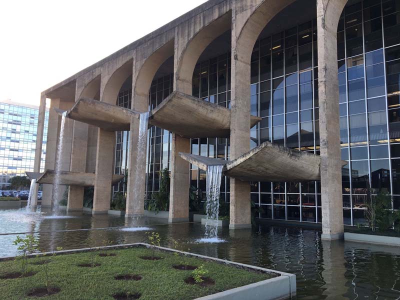 A modernist building with large concrete arches and cascading water features flowing from protruding slabs into a reflective pool, surrounded by greenery.