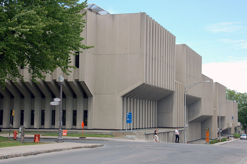 A concrete building with geometric, angular shapes and deep-set vertical grooves is seen from the street. Inside the grooves are tall slim windows.