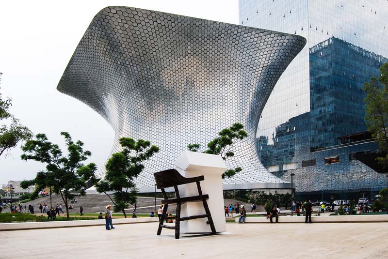 A large, silver-colored abstract building with hexagon shapes sits next to a taller building with glass windows.